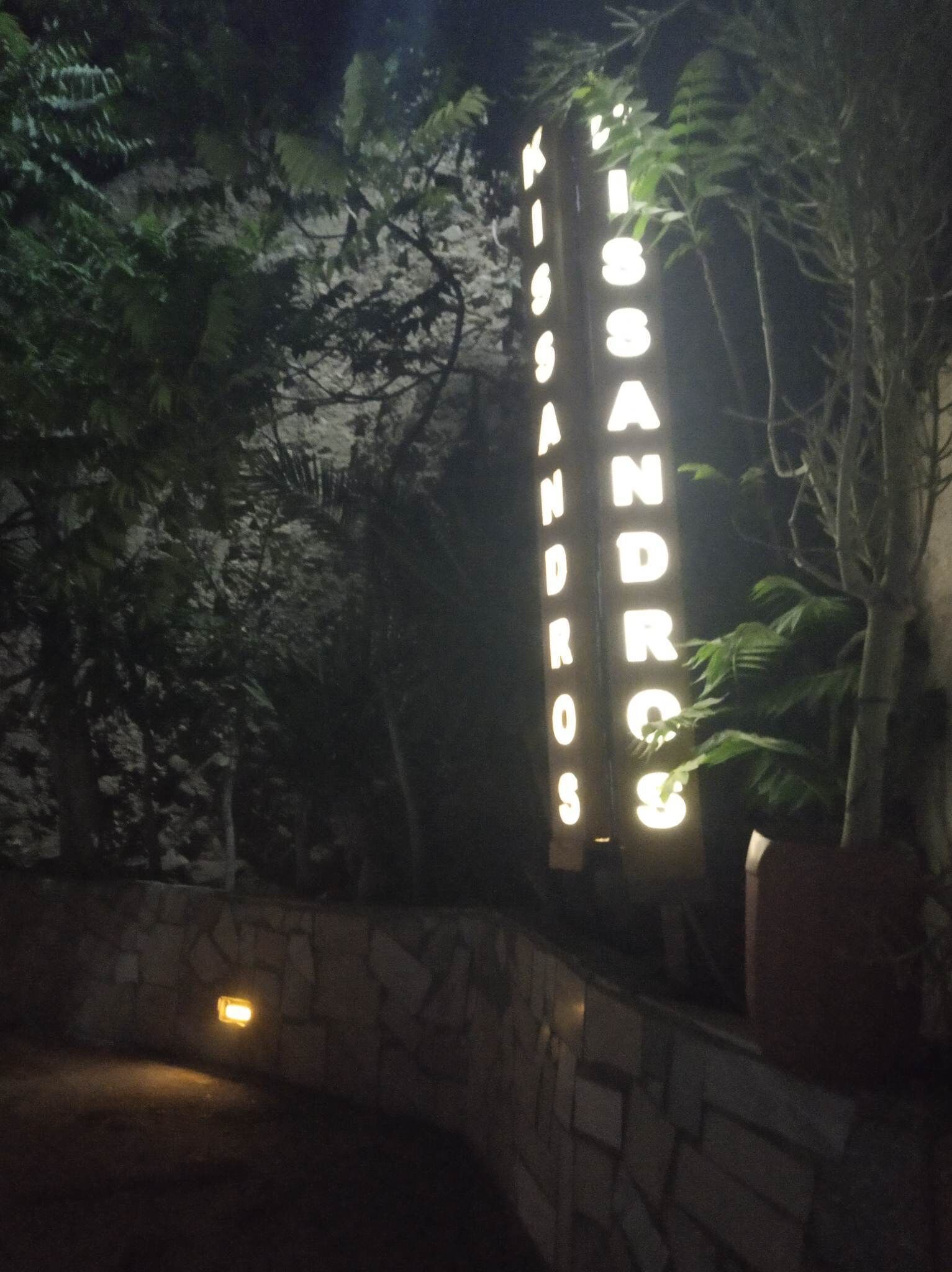 Uma placa com o nome "MIGUEL ANDROS" iluminada à noite, rodeada por folhagem.
