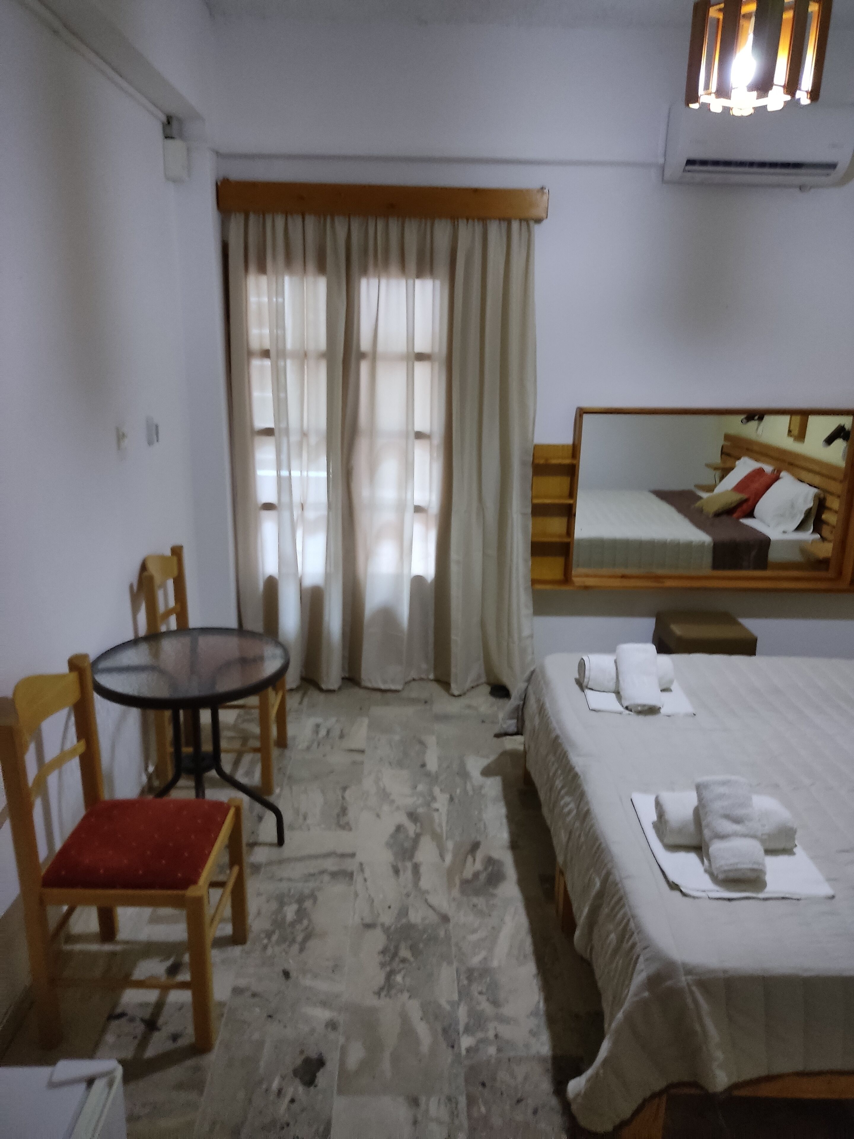 Une chambre d’hôtel avec un lit, une petite table, une chaise, une fenêtre avec des rideaux et un miroir au mur.