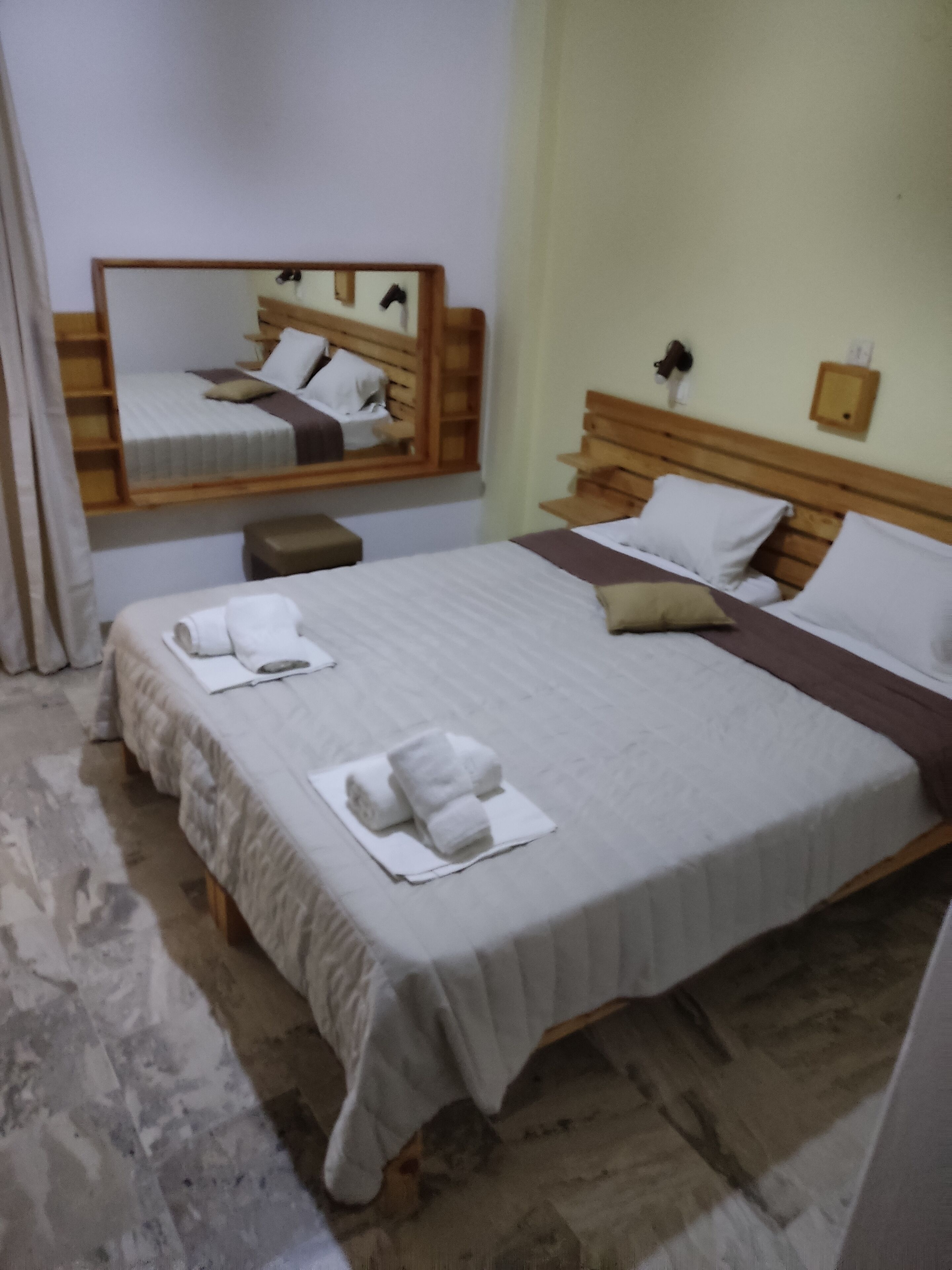 A hotel room with a large bed, a wooden headboard, a bedside table, a mirror, and a small bench.