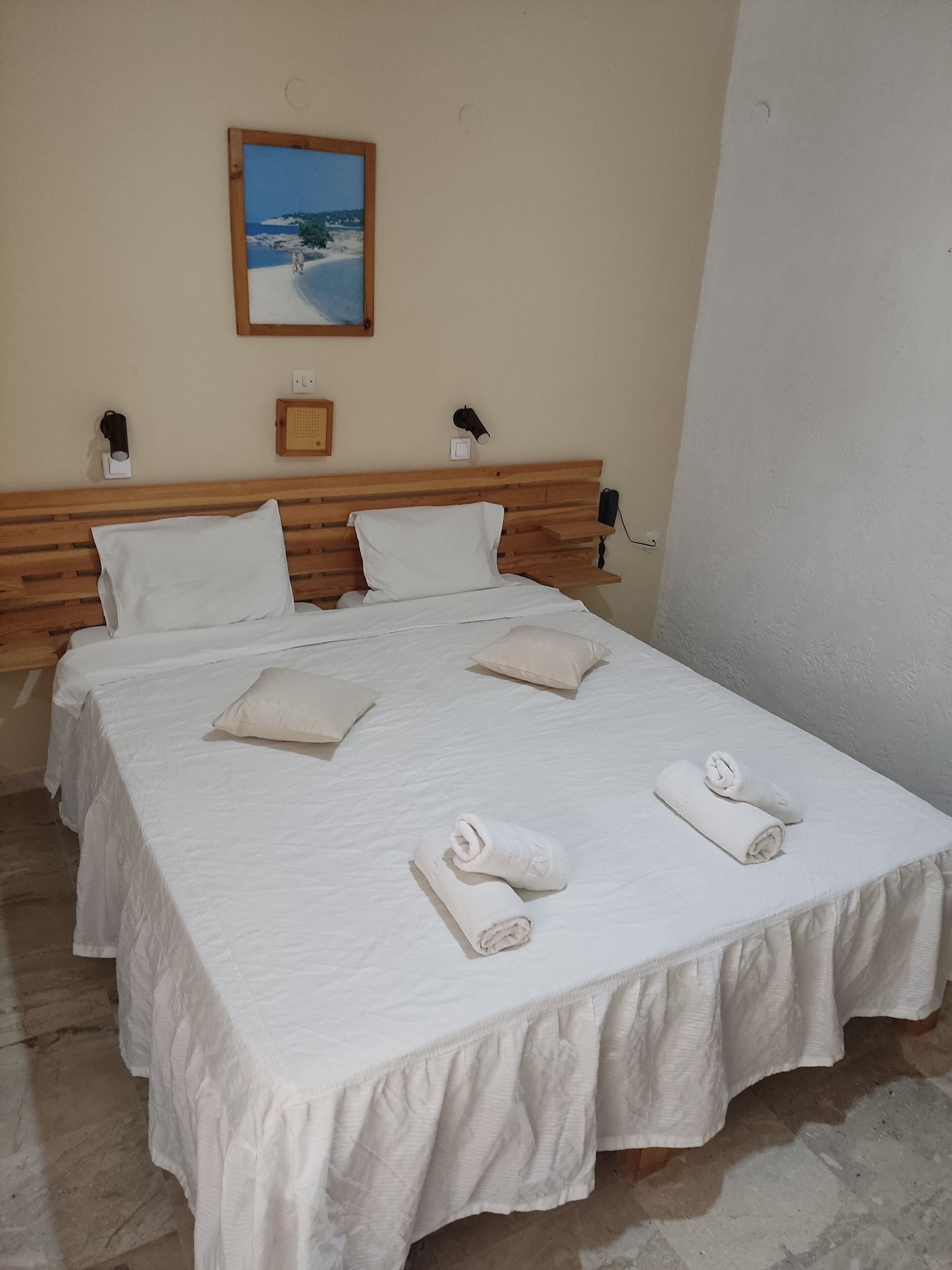 Una cama bien hecha con sábanas y almohadas blancas, un cabecero de madera y un cuadro de una playa en la pared.
