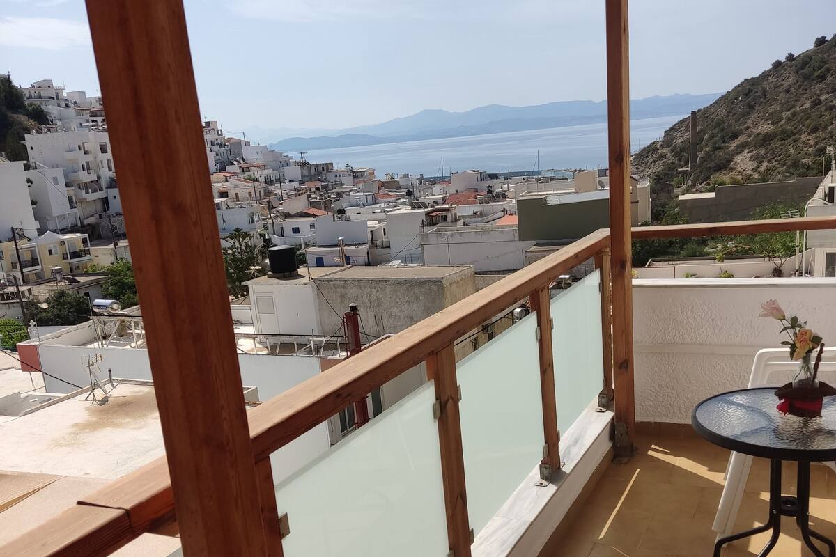 Un balcón con vistas a un pueblo costero, barandillas de madera y una pequeña mesa con un florero.