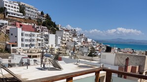 A balcony with a glass table and chairs overlooking a coastal town with white buildings and a view of the sea.
