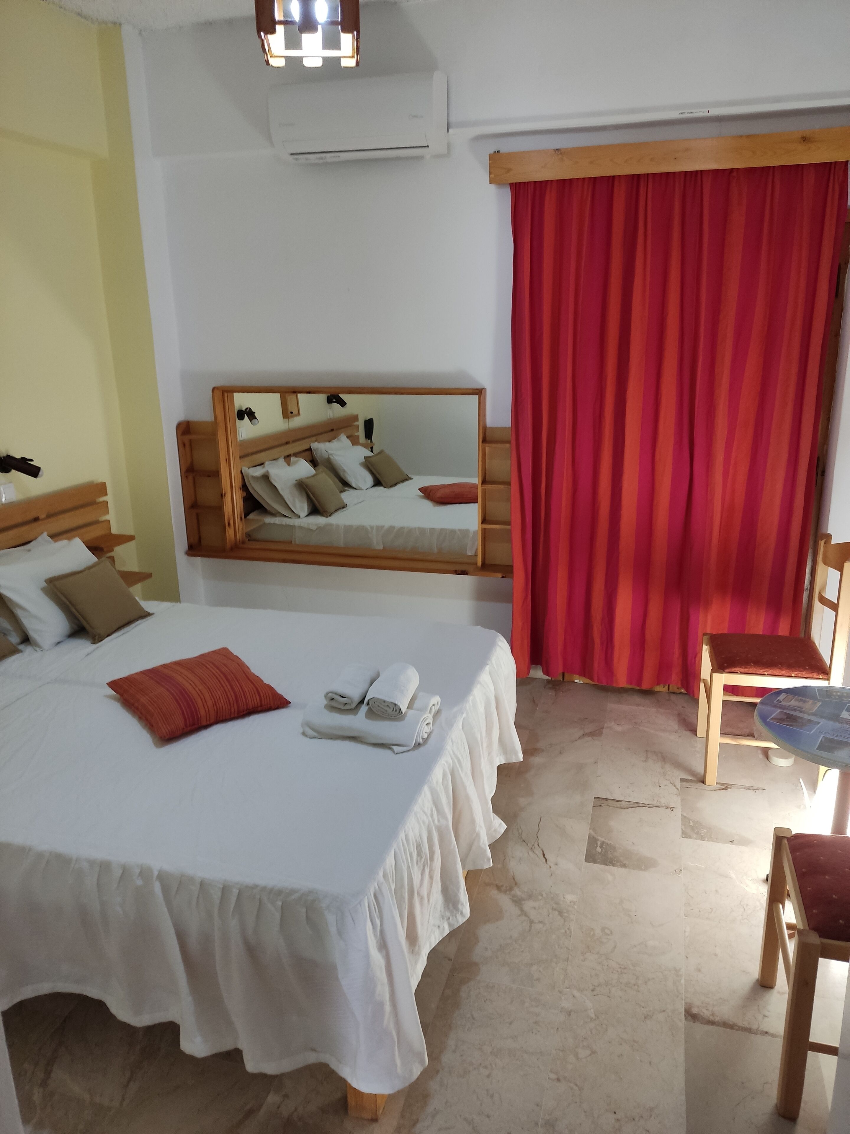A hotel room with a large bed, a wooden headboard, a mirror, a red curtain, a small round table, and a chair.