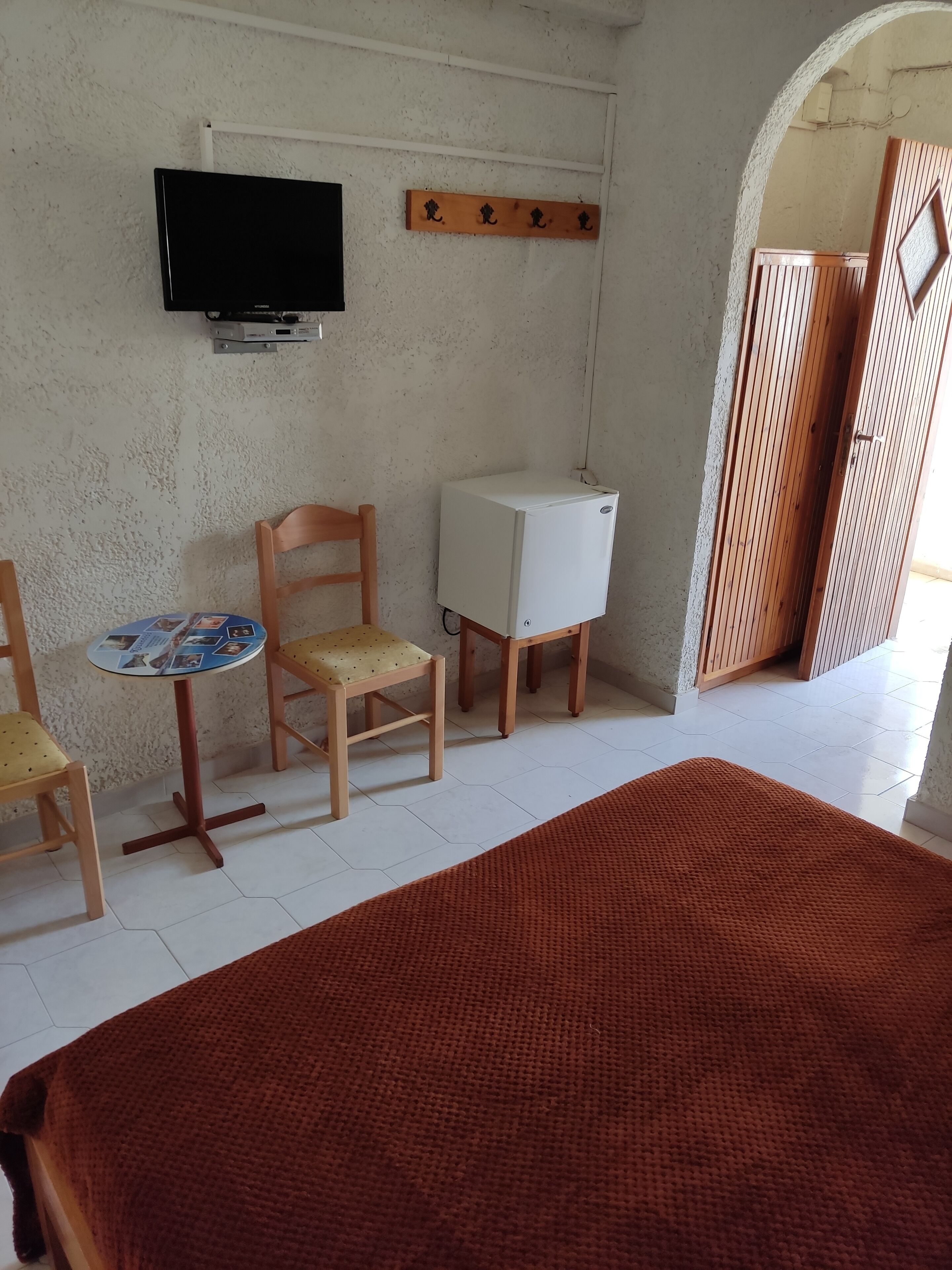 Una habitació amb llit, taula petita amb cadires, televisió penjada a la paret i un armari de fusta.
