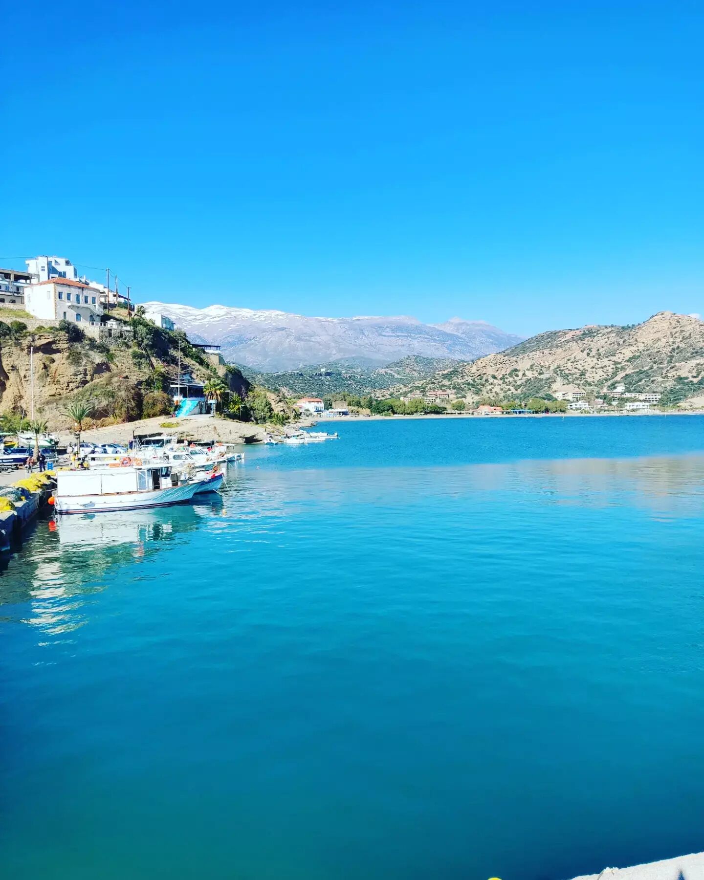 A marina with boats docked, a hillside with buildings, and a clear blue lake under a sunny sky.