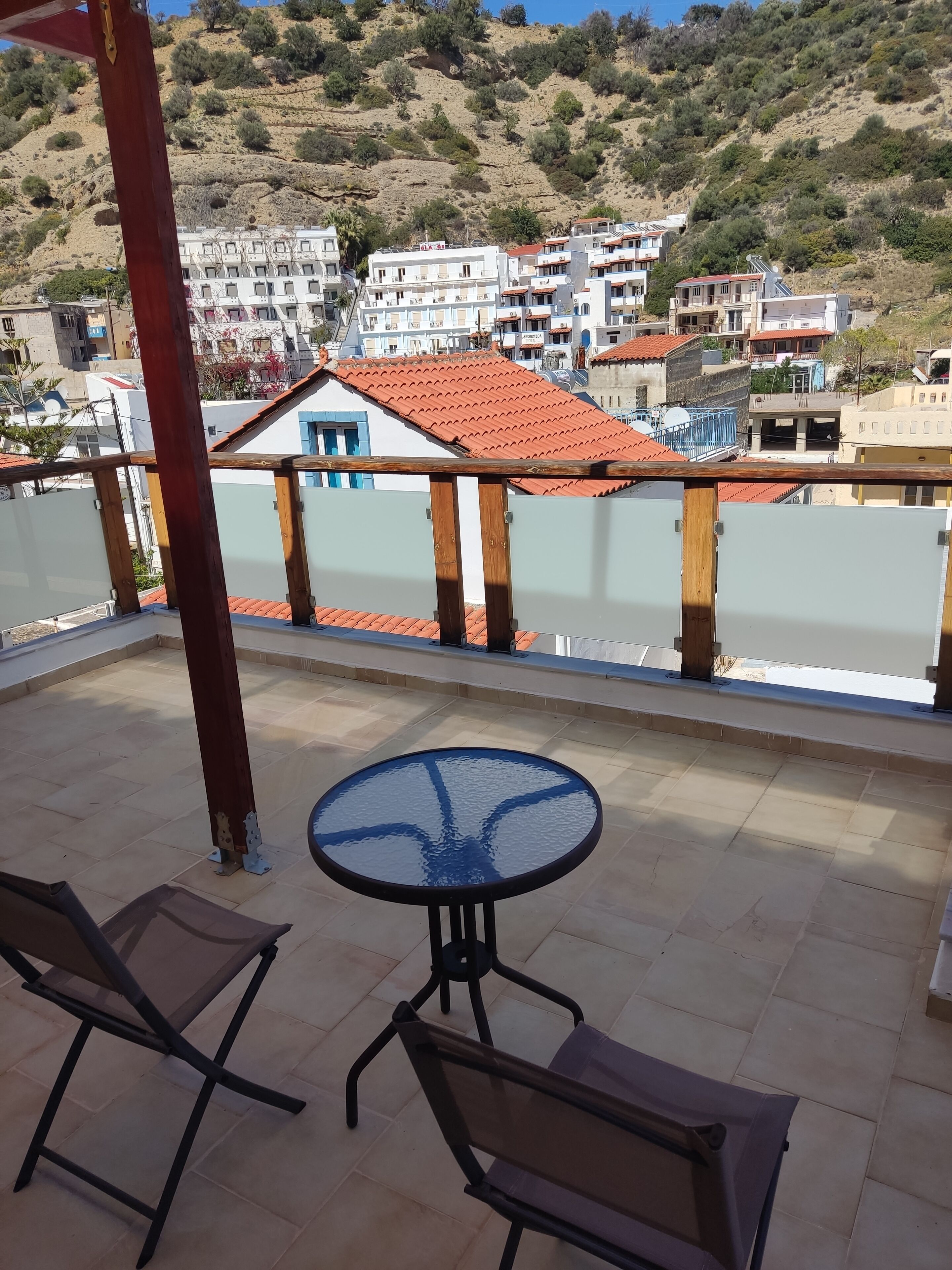 Un balcon doté d’une table ronde en verre et de deux chaises, offrant une vue sur une colline parsemée de bâtiments.