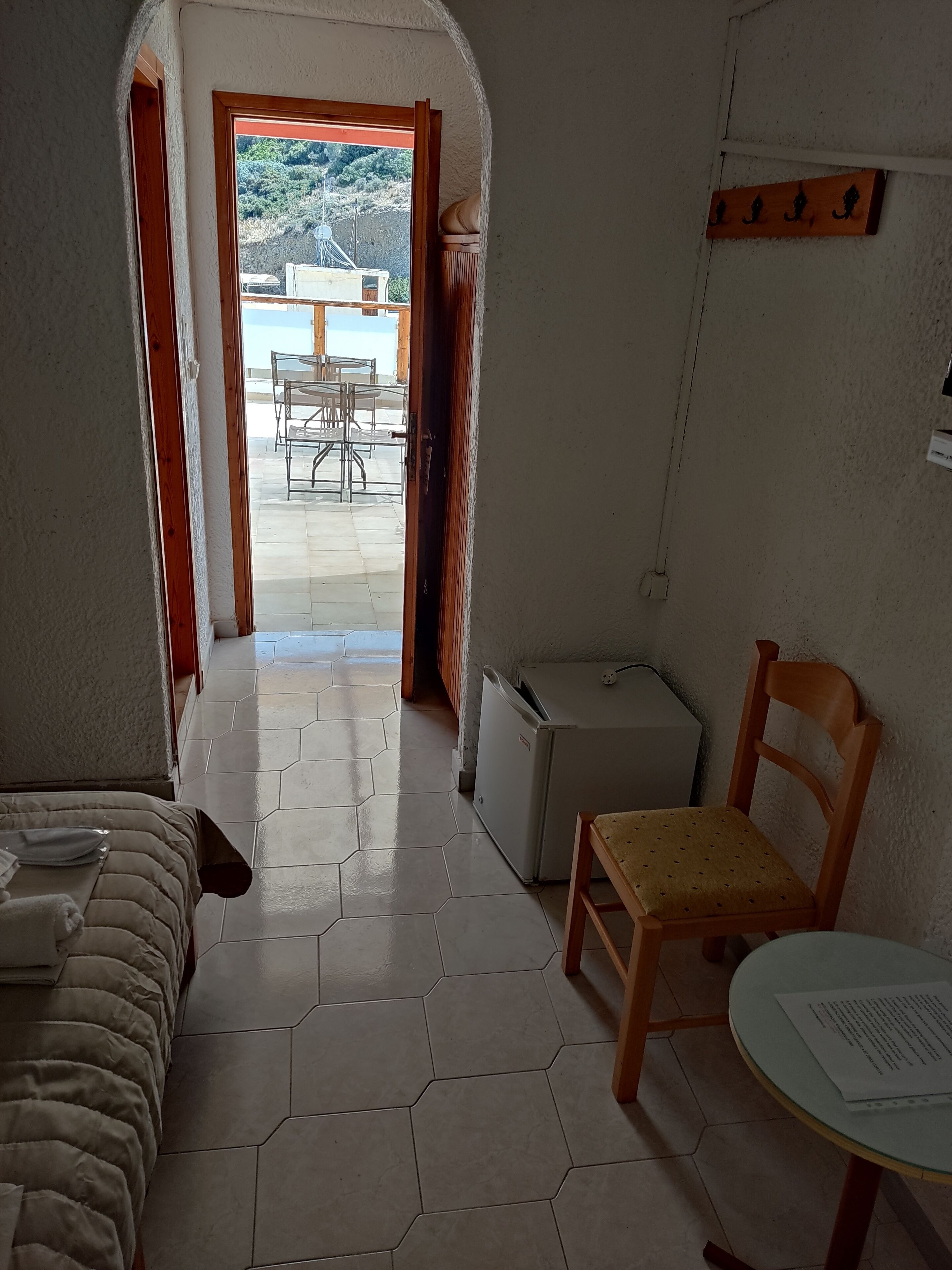 Una habitación con cama, una silla, una mesita y un refrigerador. Hay una puerta que da a un espacio exterior con una mesa y sillas.