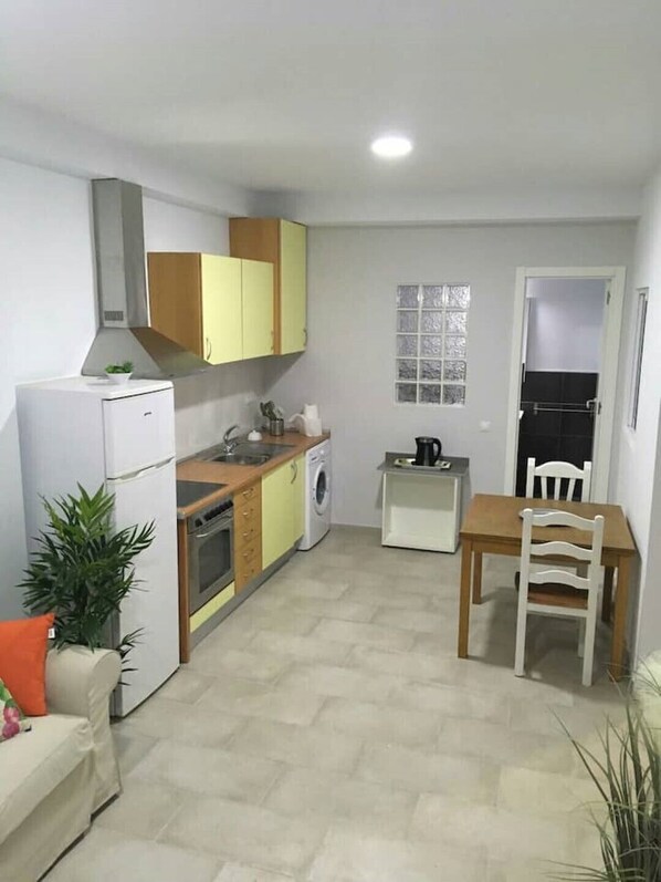 A compact kitchen with a refrigerator, oven, microwave, and washing machine. There is a dining table with a chair and a small window above it.