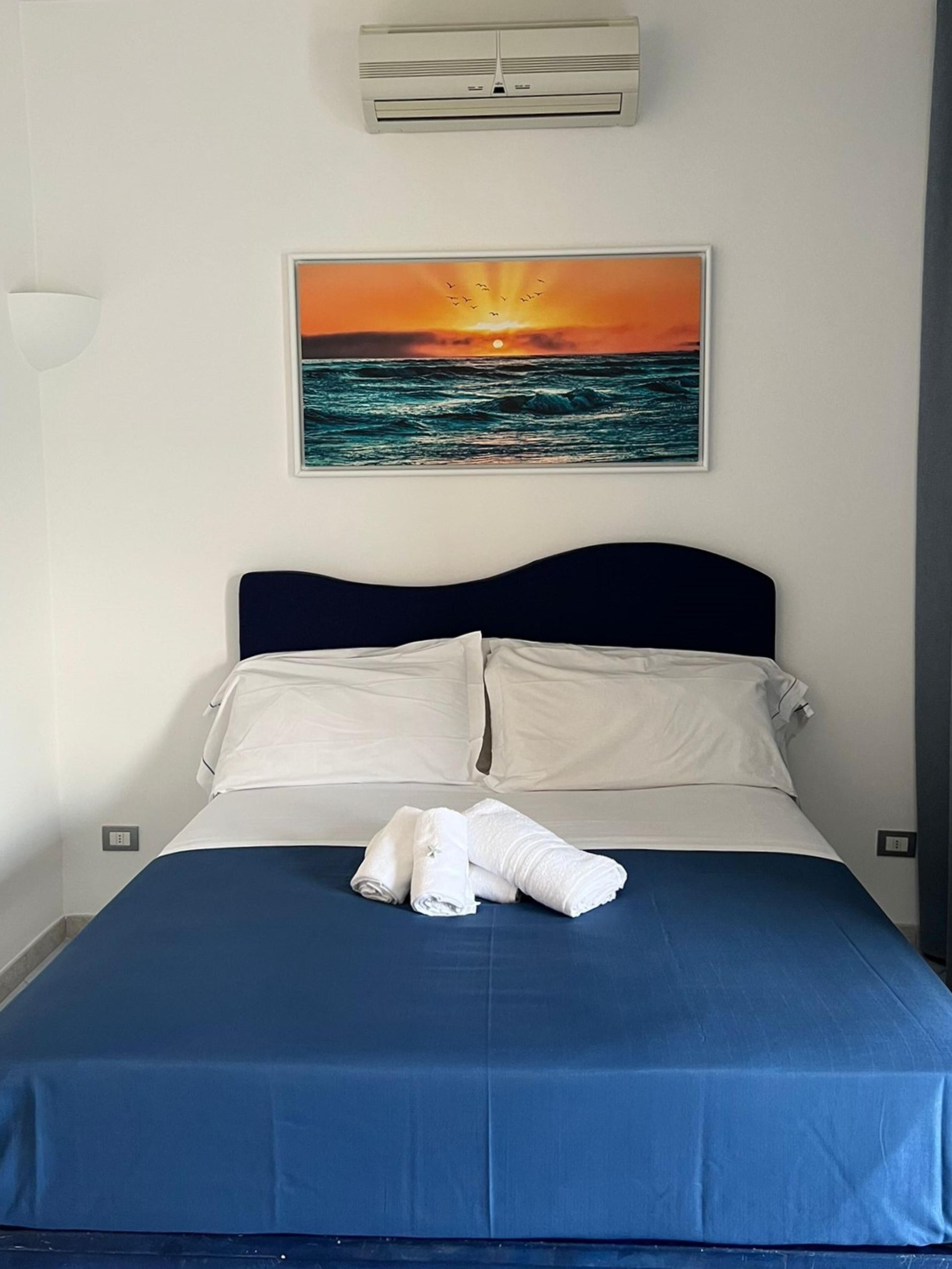 Una cama bien hecha con una colcha azul y almohadas blancas, un cuadro de un atardecer sobre el océano en la pared, y un equipo de aire acondicionado blanco sobre la cama.