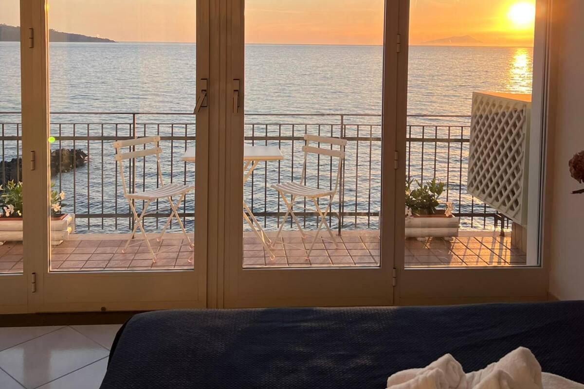 Een kamer met uitzicht op zee tijdens zonsondergang, een balkon met stoelen en een bed met een handdoek.