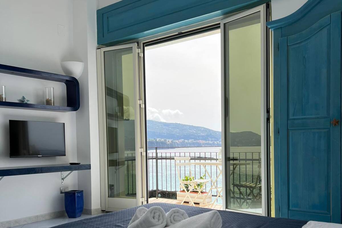 Een hotelkamer met een bed, een televisie, een balkon met uitzicht op zee, en een blauwe deur.