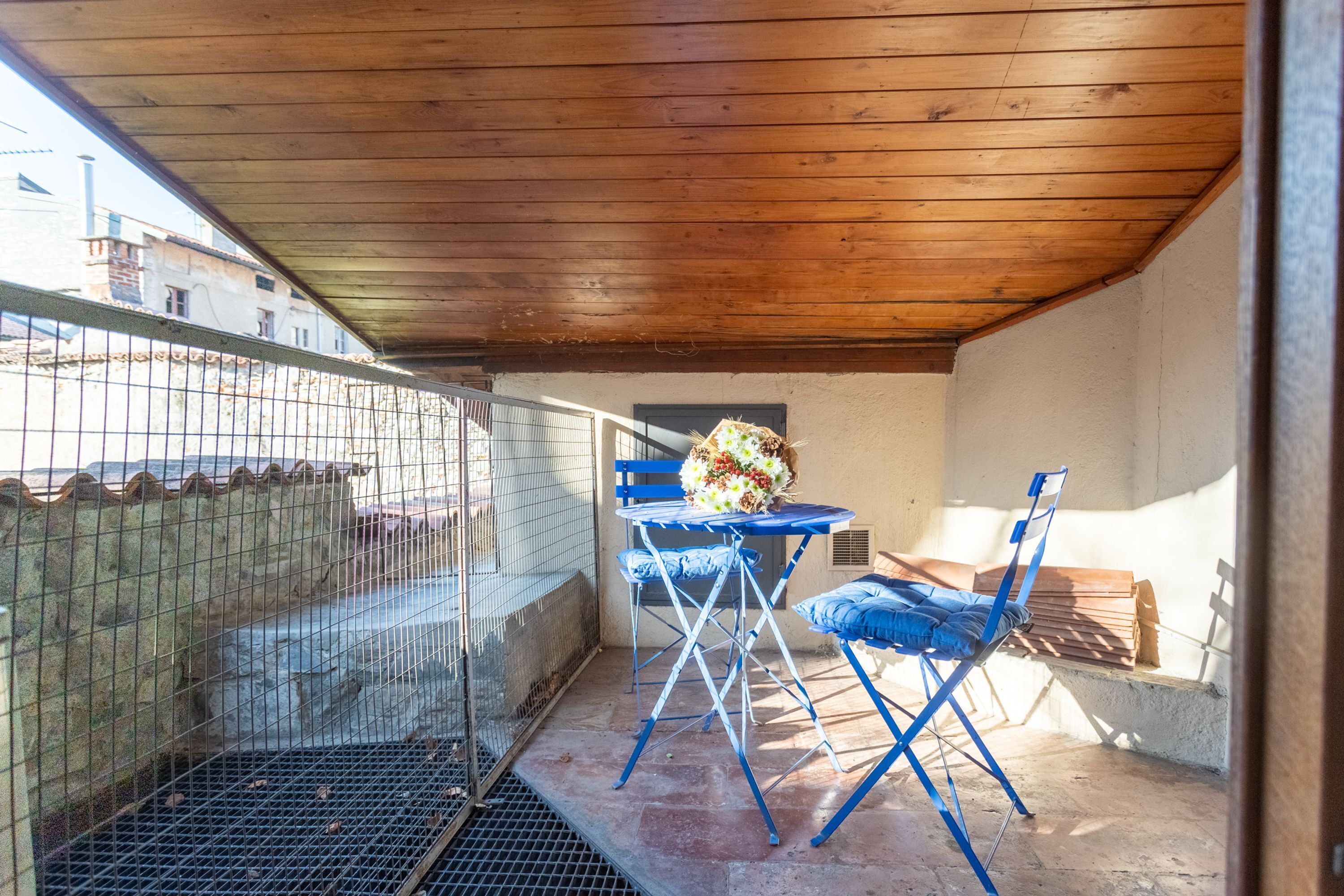A covered outdoor seating area with blue chairs and a table, a metal fence, and a view of buildings in the background.
