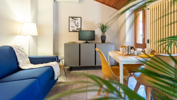 A cozy living room with a blue sofa, a small table with a lamp, a TV on a cabinet, and a dining area with yellow chairs. - San Fedele Apartments (Como)