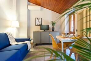 A cozy living room with a blue sofa, a small table with a lamp, a TV on a cabinet, and a dining area with yellow chairs. - San Fedele Apartments (Como)