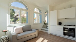 A modern kitchen with white cabinets and a built-in oven. There is a grey sofa with a cushion and a small side table with a vase of flowers. Large windows provide natural light and a view of the outside. - Mezzegra Apartments in Tremezzo (Tremezzina)
