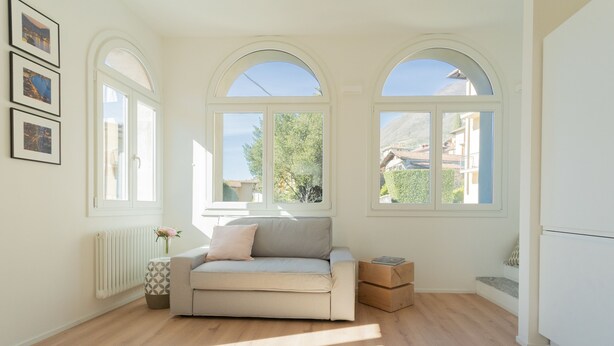 A spacious room with a beige sofa, wooden flooring, and large arched windows offering a view of the outdoors. - Mezzegra Apartments in Tremezzo (Tremezzina)