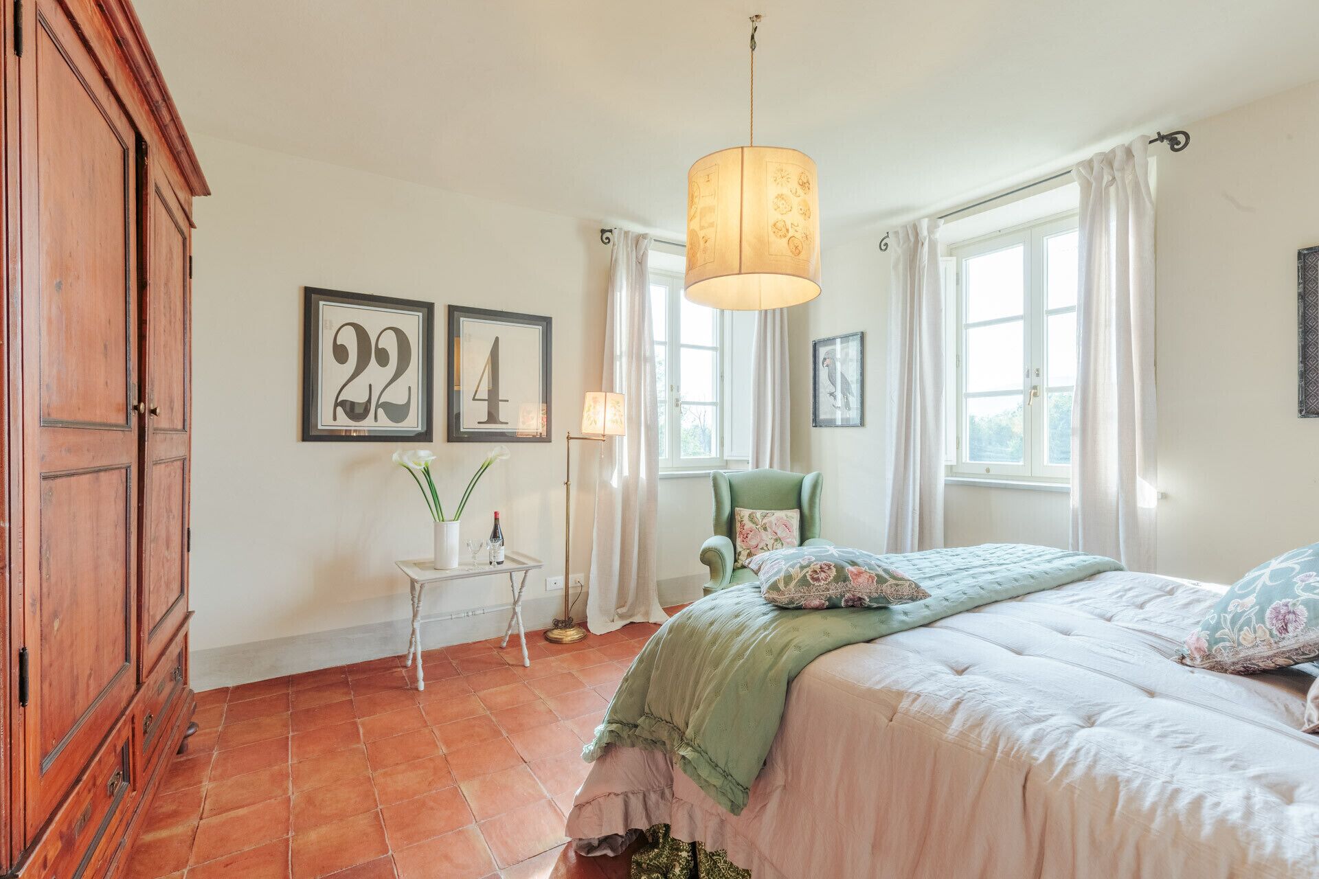 Un dormitorio con una cama, una silla, una mesita con un florero y una botella de vino, y dos números enmarcados en la pared.