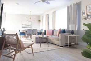 Een moderne woonkamer met een bank, salontafel en stoelen. Links staat een televisie en rechts een raam met gordijnen.