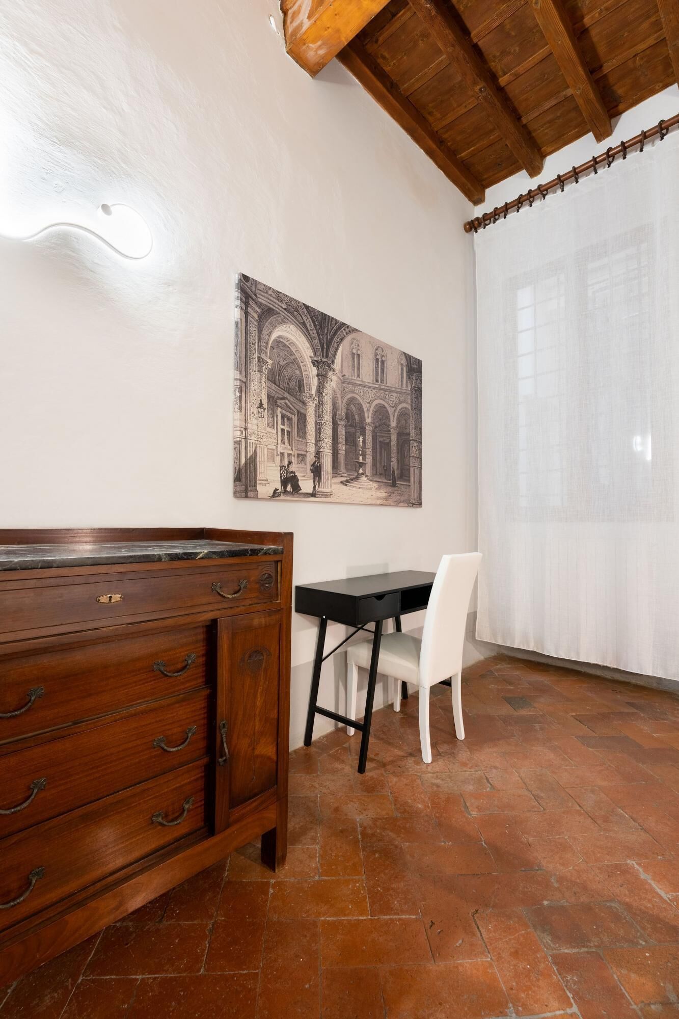 Una habitación con un aparador de madera, una mesa negra y una silla blanca. En la pared, un cuadro grande enmarca una escena arquitectónica.