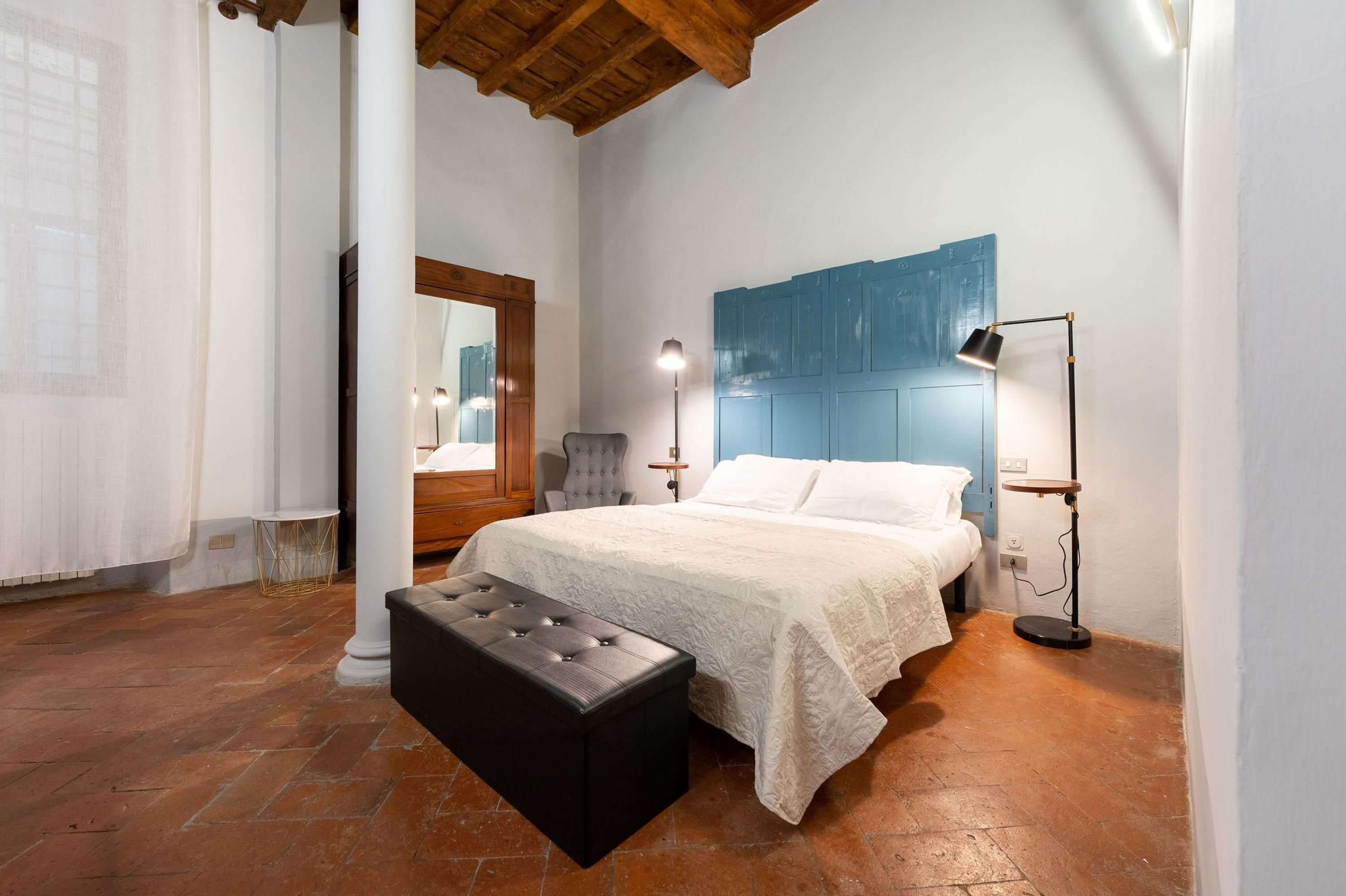 A bedroom with a bed, a nightstand, a chair, a bench, a floor lamp, and a wall-mounted mirror.