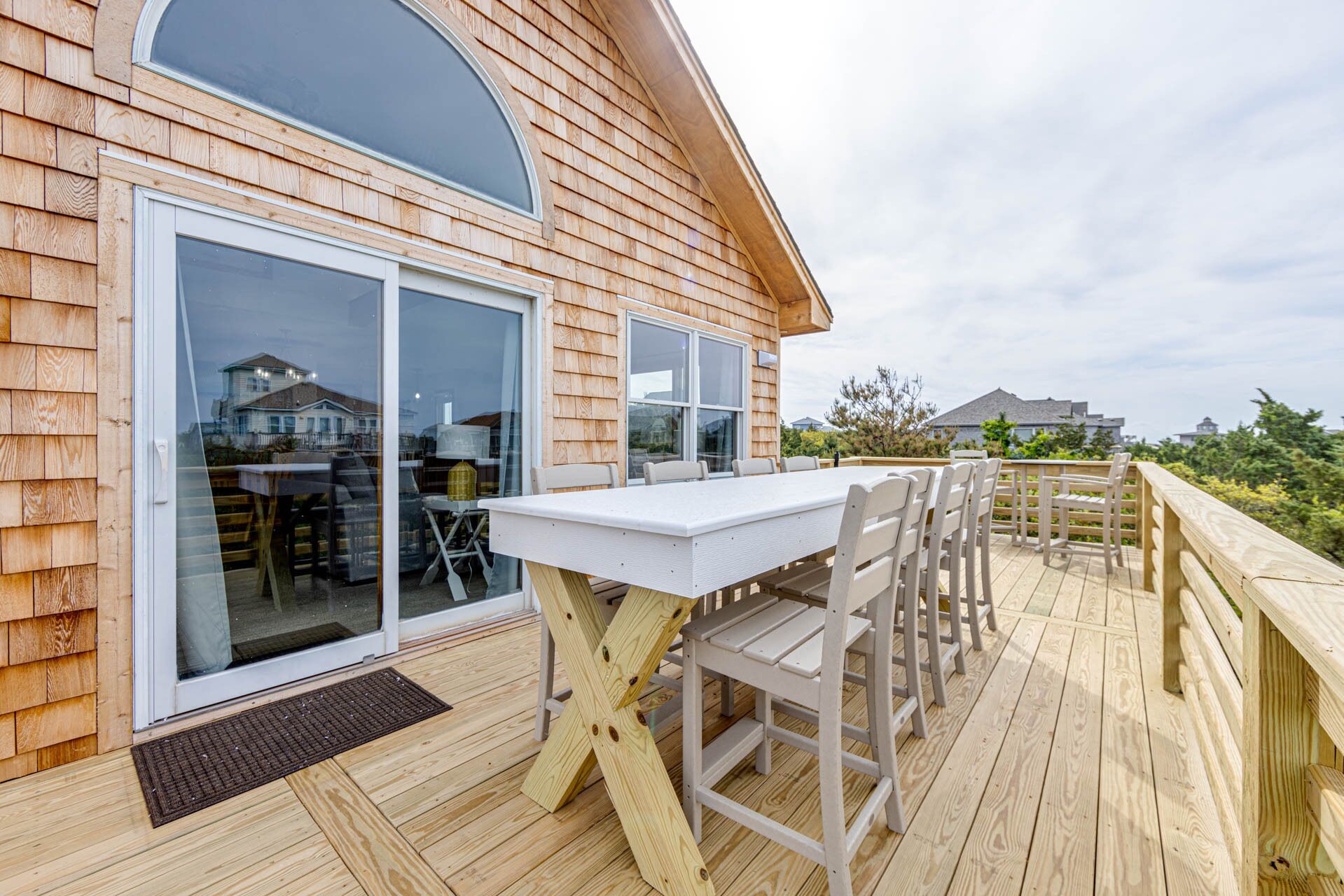 A wooden deck with a white table and chairs, large sliding glass doors, and a view of the surrounding area.
