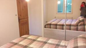 Een slaapkamer met een houten deur, een bed met een geruit plaid en een spiegel die de kamer weerkaatst.