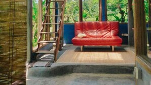 A room with a red sofa, a wooden staircase, and large windows offering a view of greenery. - Pondok Apsari 2 (Sukasada)