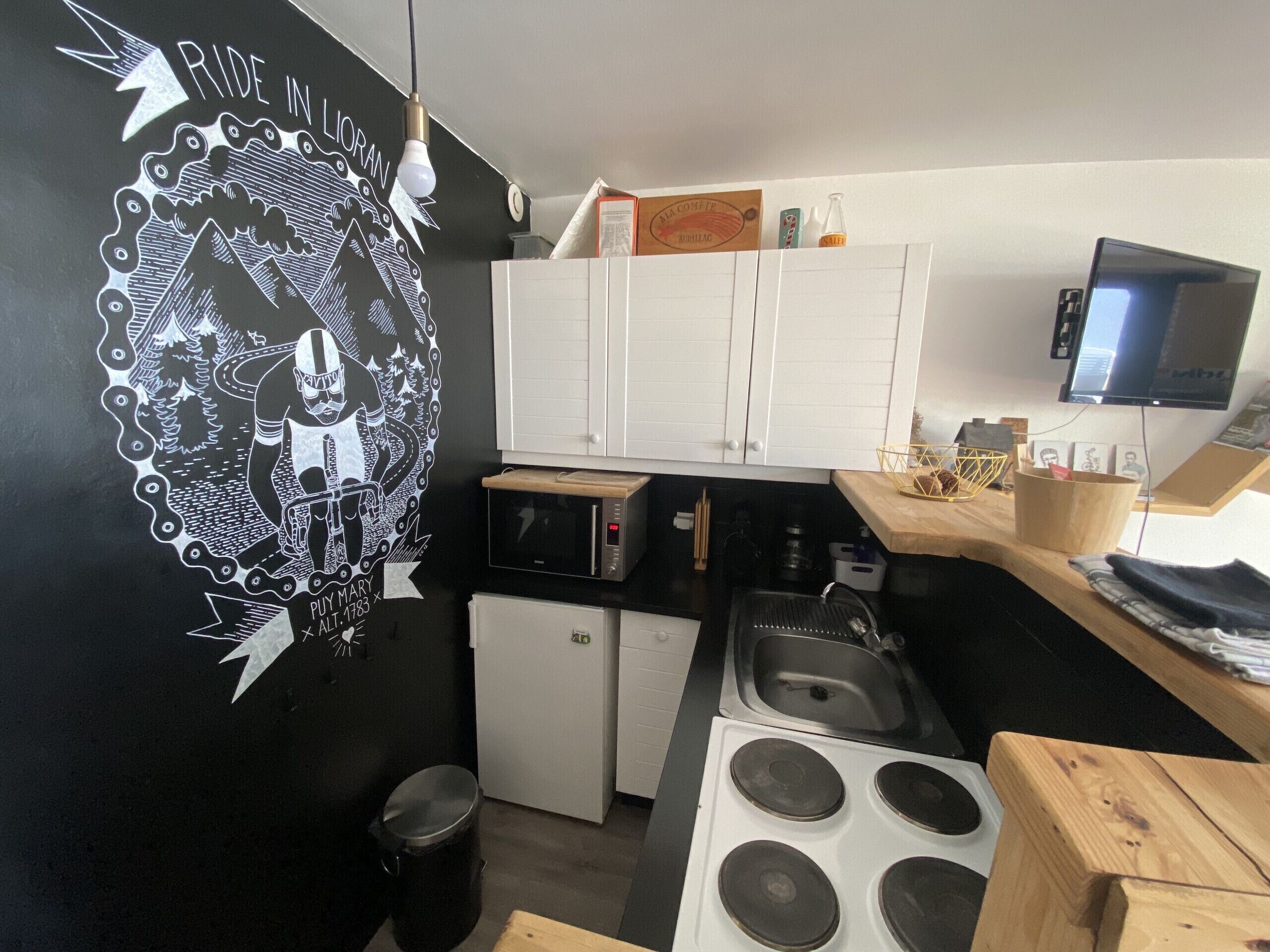 Eine kleine Küche mit weißen Schränken, einer Mikrowelle, einem Herd und einem Spülbecken. An der Wand hängt eine Dekoration und ein Fernseher.