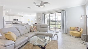 A modern living room with a sectional sofa, a glass coffee table, and a yellow armchair. There is a kitchen in the background with white cabinets and a built-in oven. - Modern Waterfront Home in Artesia | Pool/Gym (Naples)