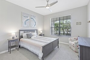 A bedroom with a bed, bedside tables, a ceiling fan, a window with blinds, and a decorative sign.