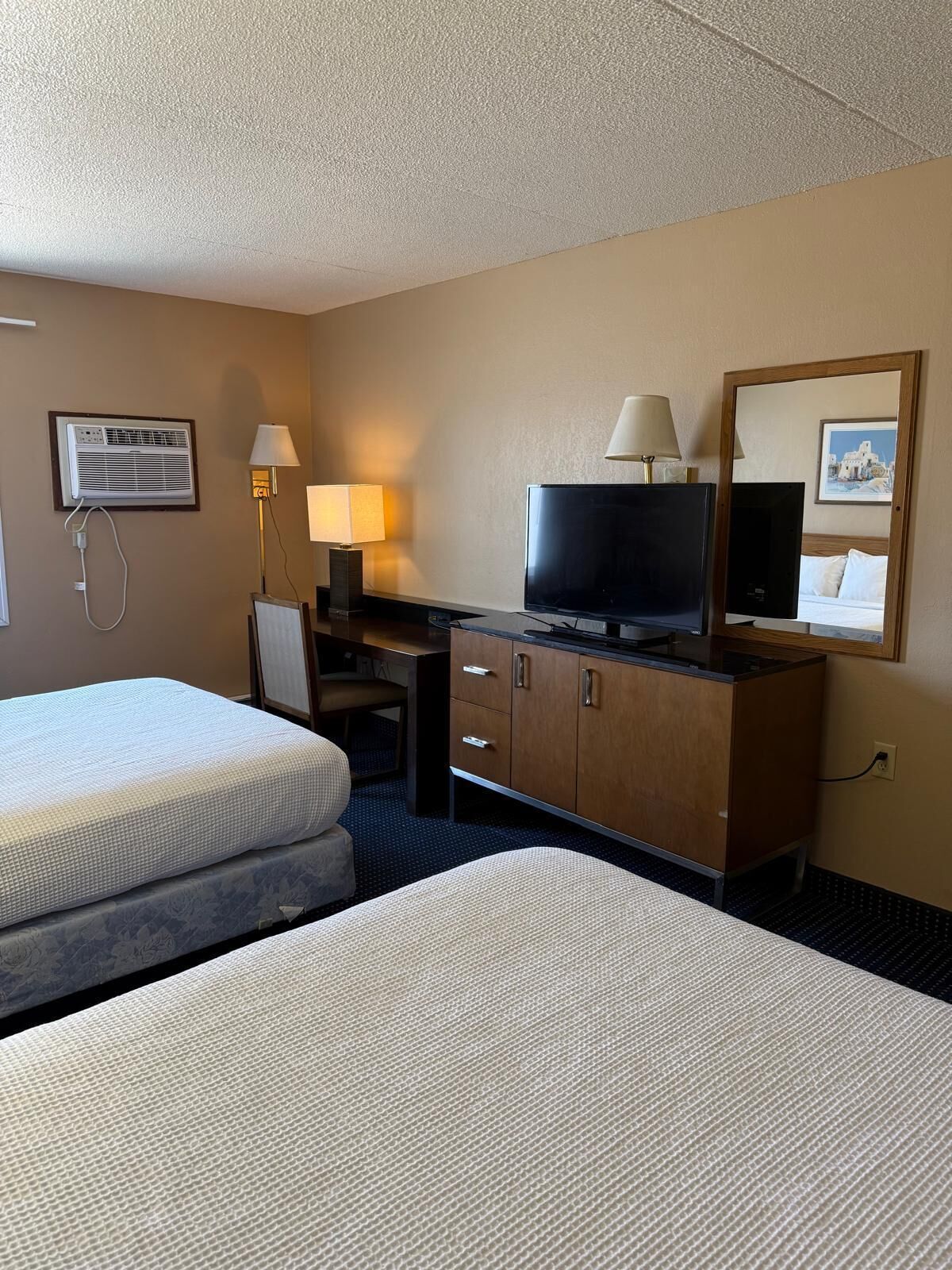 A hotel room with two beds, a desk with a television, a mirror, and a wall-mounted air conditioner.