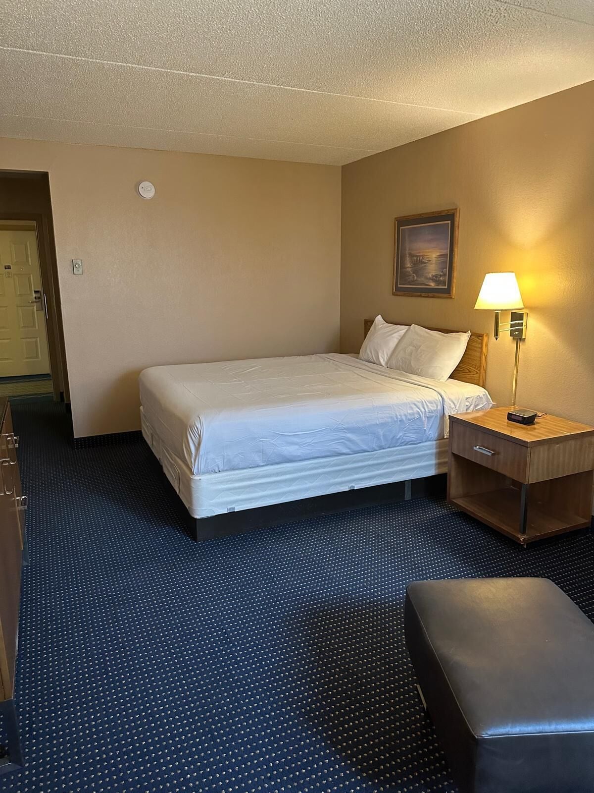 Ein Hotelzimmer mit Bett, Nachttisch mit Lampe, Bank und einer Tür zu einem weiteren Raum.