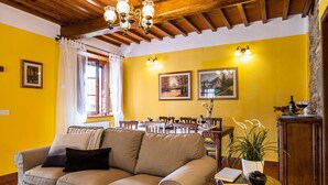 Ein gemütliches Wohnzimmer mit einer Couch, einem Esstisch und Stühlen. An den Wänden hängen Bilder in Rahmen und über dem Tisch hängt ein Kronleuchter.