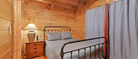A wooden cabin room with a metal bed, a bedside table with a lamp, and a curtain.