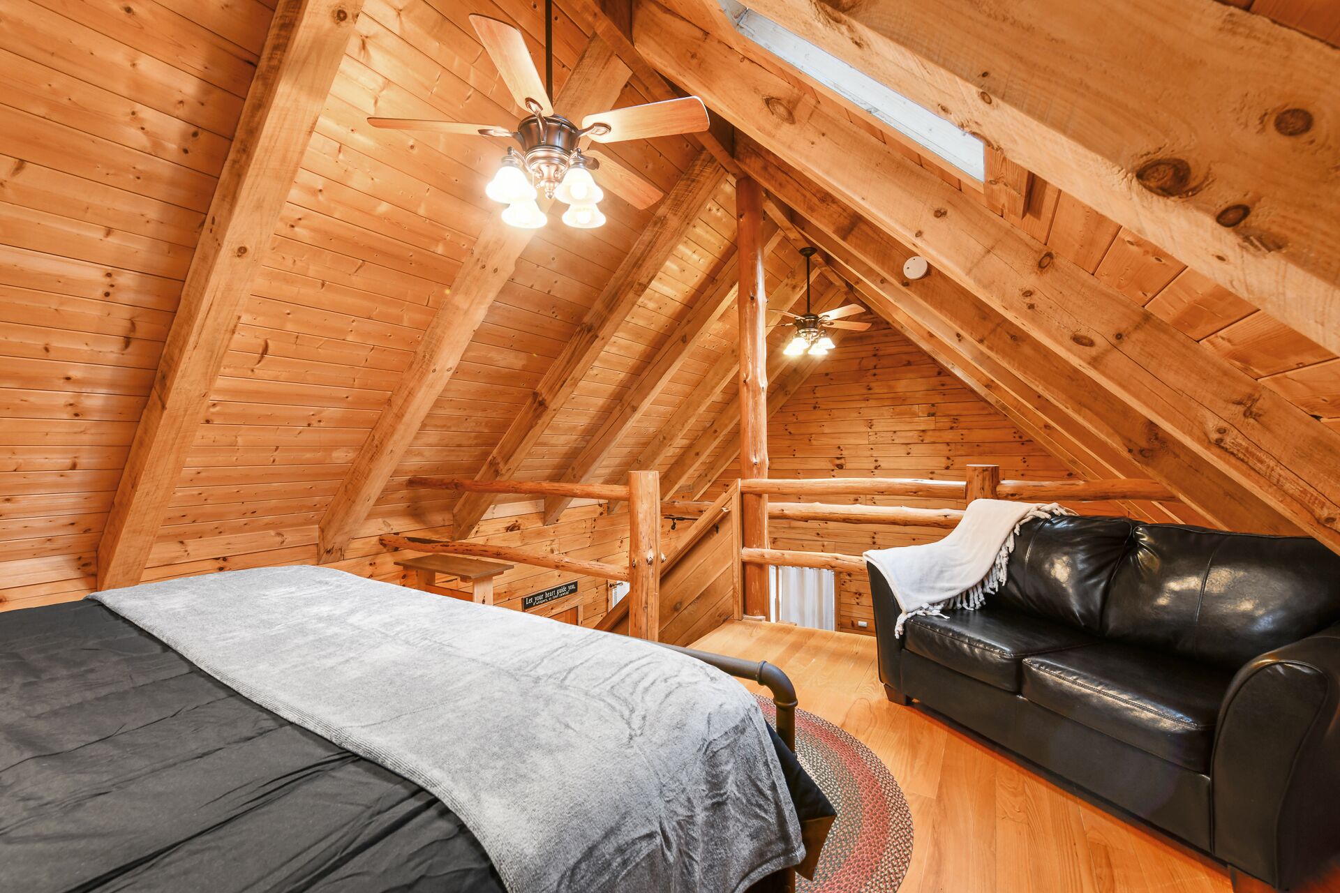 A cozy attic bedroom with a wooden ceiling, a bed, a black leather sofa, and a ceiling fan.