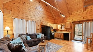 A cozy cabin interior with a wooden ceiling, a living room with a sofa and cushions, a dining area, and a kitchen with wooden cabinets and appliances. - Cabin In Hocking Hills! Pet Friendly, Hot Tub, Secluded. (Logan)