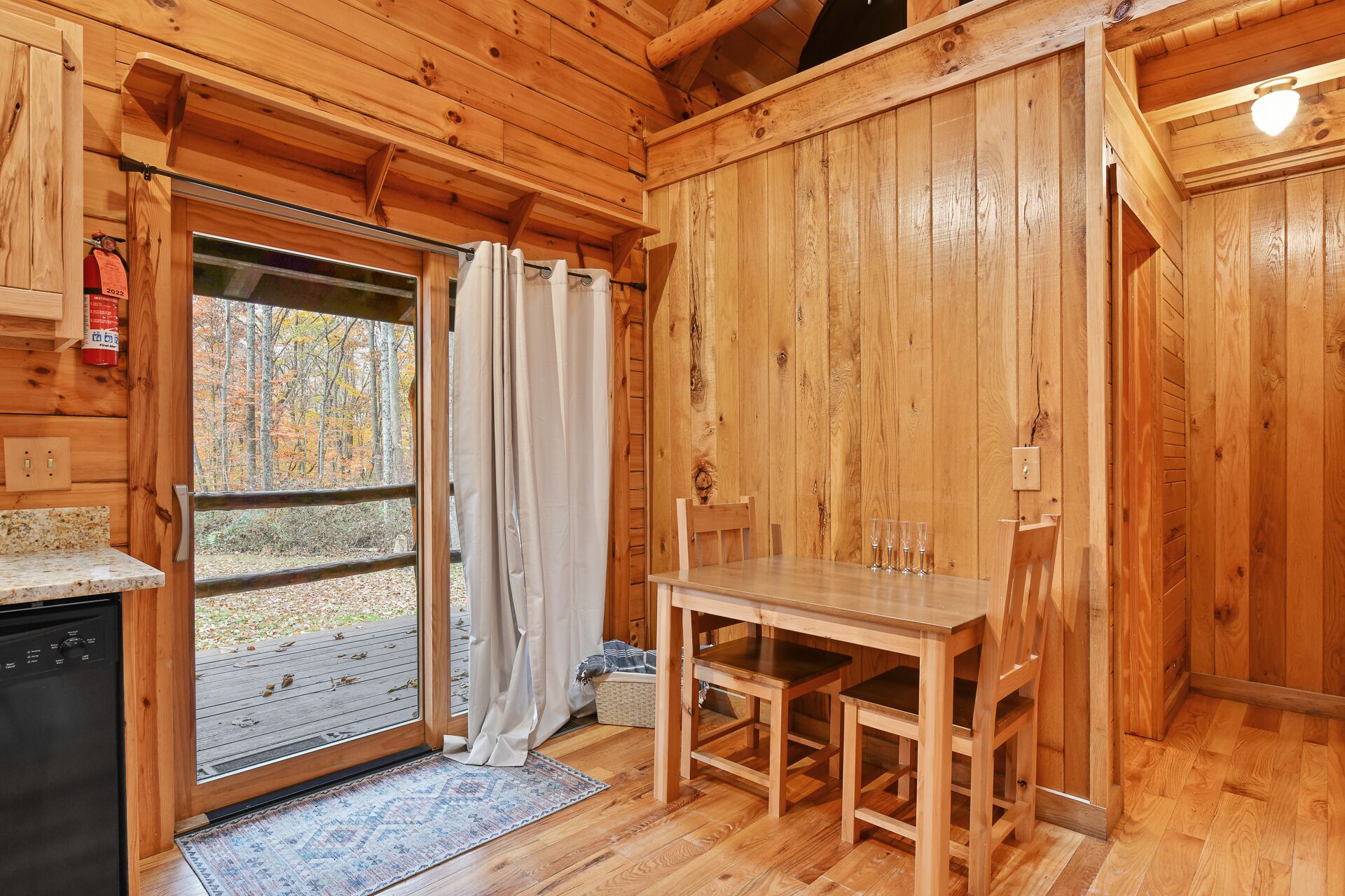 A wooden cabin interior with a dining table, chairs, a window with curtains, and a view of a forest.