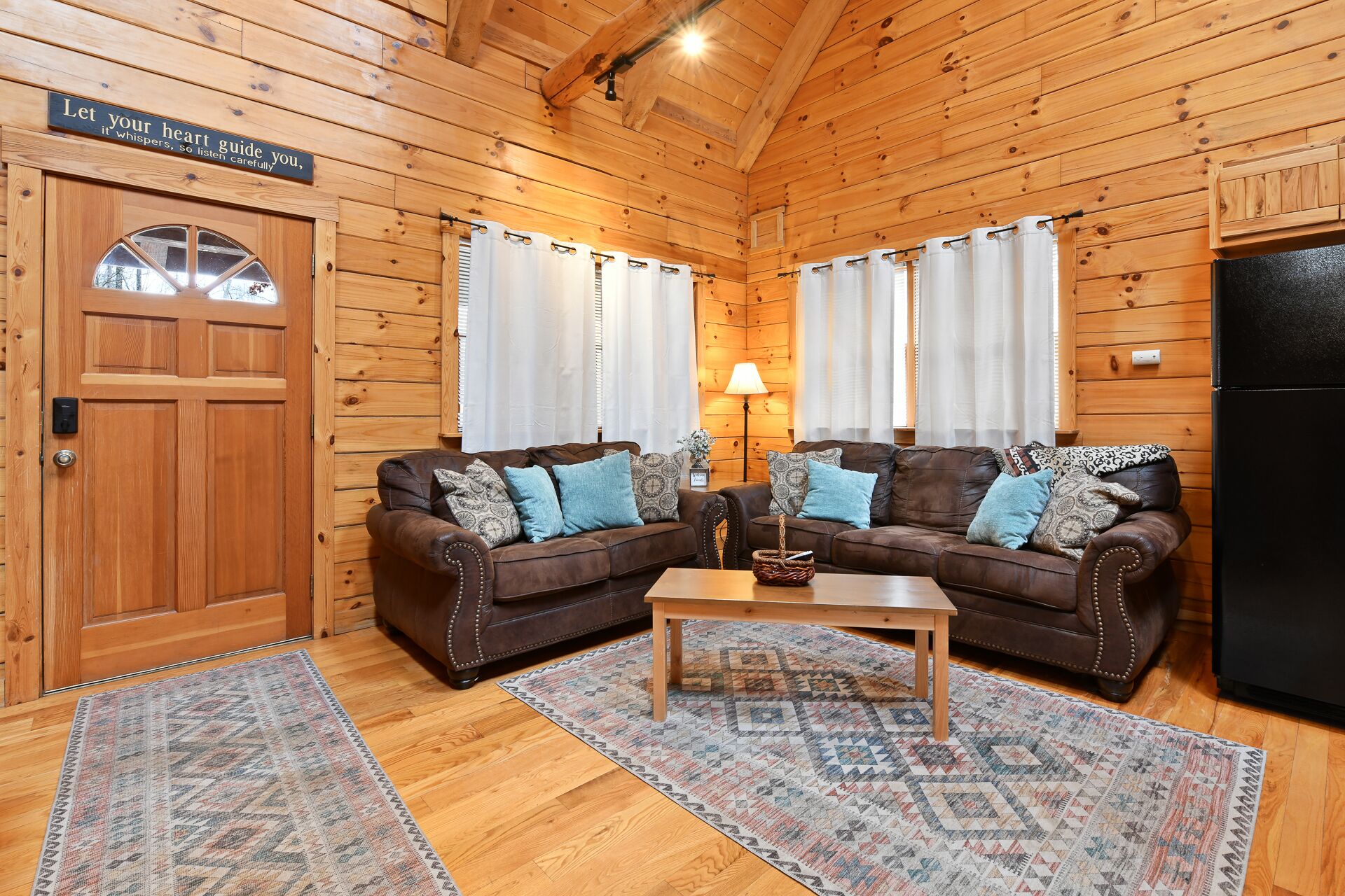Interior acogedor de una cabaña con paredes de madera, dos sofás de cuero marrón con cojines estampados, una mesa de centro de madera y un refrigerador negro.