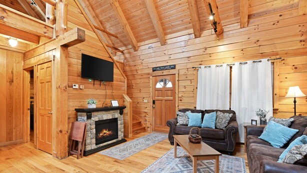 A cozy log cabin interior with a stone fireplace, wooden walls, a flat-screen TV, and a comfortable seating area with a sofa and armchairs. - Cabin In Hocking Hills! Pet Friendly, Hot Tub, Secluded. (Logan)