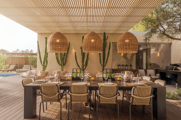 Un área de comedor al aire libre con una mesa preparada para cuatro personas, rodeada de sillas, bajo un toldo con lámparas colgantes y cactus al fondo.