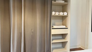 Habitación con armario que incluye varios estantes y espacio para colgar, cama con edredón blanco y cortina entreabierta.