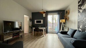 Una sala de estar moderna con televisor de pantalla plana, un sofá azul oscuro, una mesa de comedor con sillas y un ventanal con vistas al exterior.
