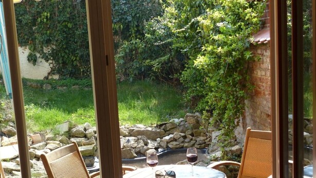 Une terrasse extérieure avec une table ronde et deux chaises en osier, un verre de vin rouge et une paire de lunettes de soleil posées sur la table.