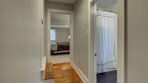 Un pasillo estrecho con un banco de madera, una puerta que da a un dormitorio con cama y ventana, y una zona de ducha con cortina.