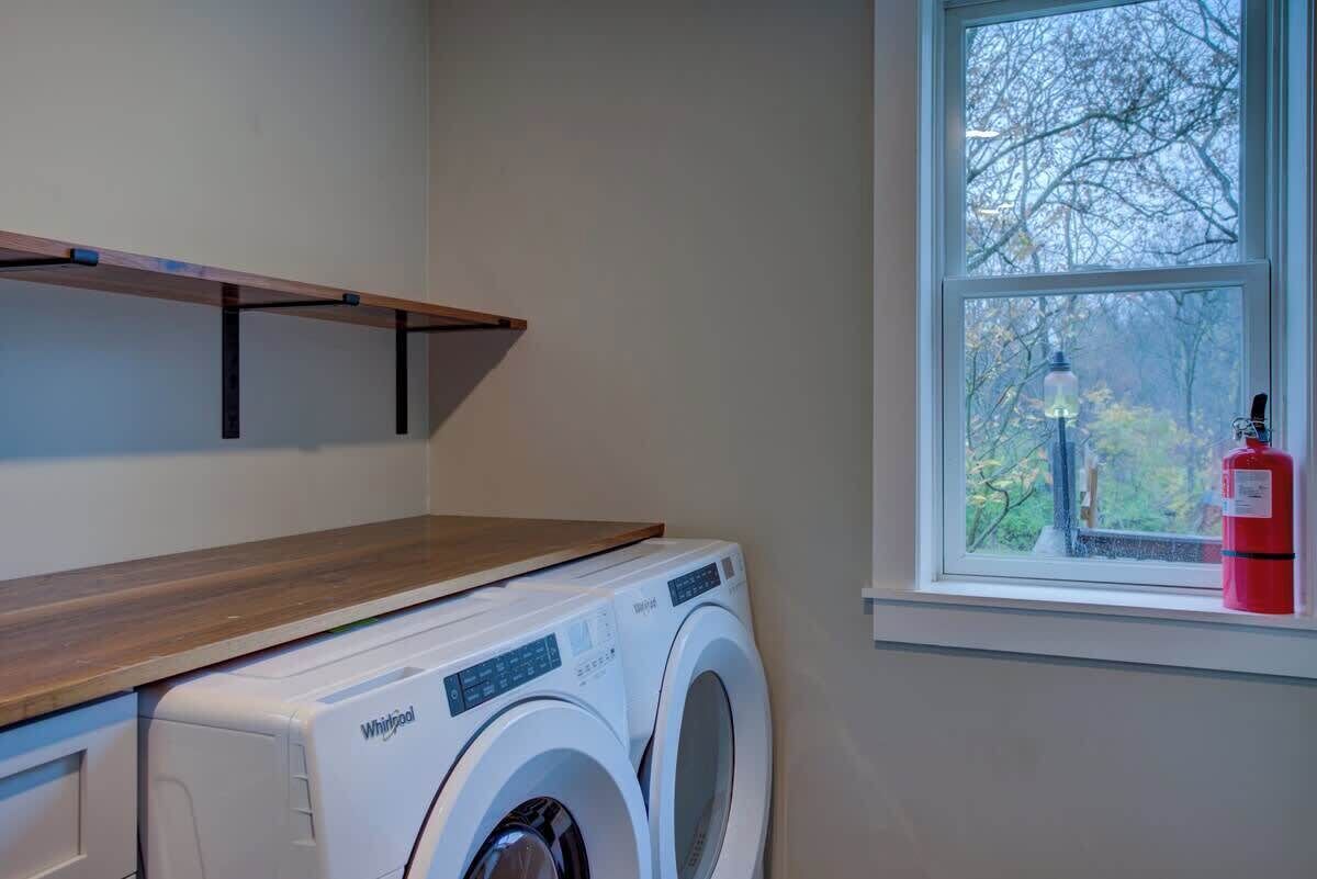 Un cuarto de lavado con lavadora y secadora Whirlpool, una ventana con vista a árboles y un extintor en el alféizar.