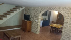 A living room with a wooden bench, a TV on a wooden cabinet, a dining table with chairs, and a kitchen area with a stone wall and a staircase. - 🌿 Gîte "Le Noyer" - Le Mas des Charrettes (Provence-Alpes)