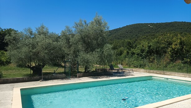A rectangular swimming pool with a clear blue water surface, surrounded by a concrete deck. There are two lounge chairs and a small table under a pergola. The pool is enclosed by a low fence, and there are trees and a hill in the background. - 🌿 Gîte "Le Noyer" - Le Mas des Charrettes (Provence-Alpes)