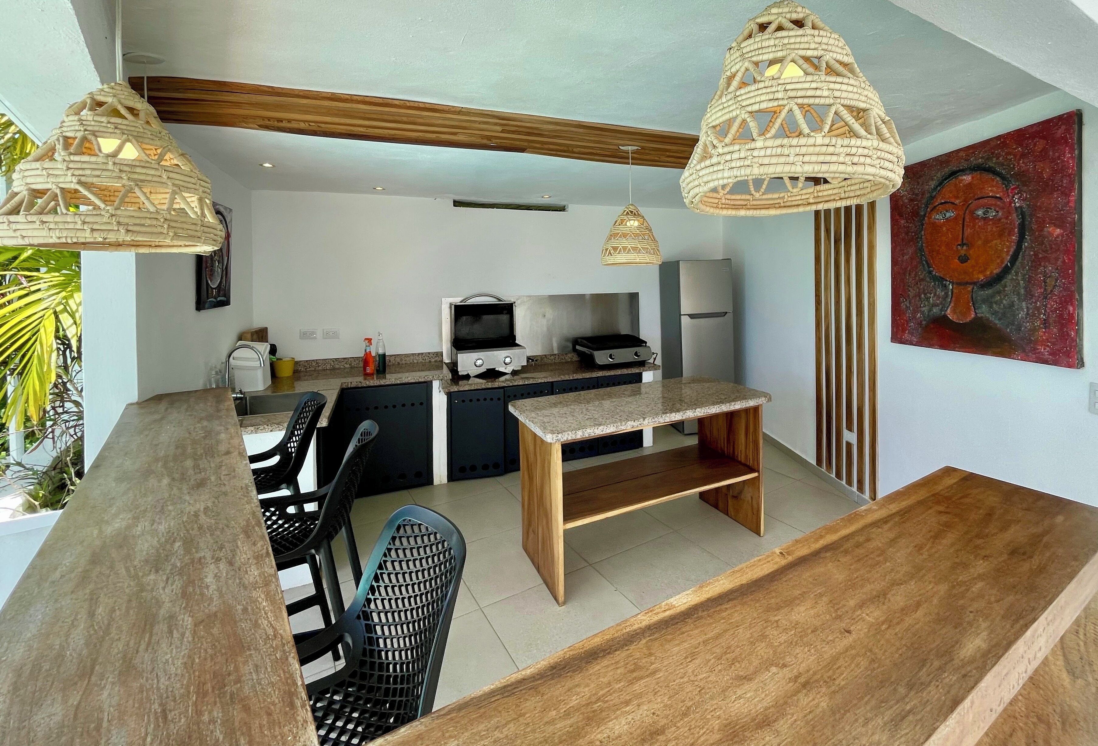 A modern kitchen with a dining area, two hanging wicker light fixtures, a wooden bench, and a painting on the wall.