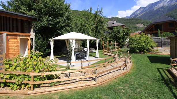 Een houten chalet met een overdekt terras, omgeven door een houten hek en weelderige begroeiing.