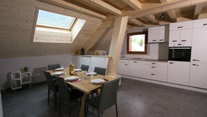 Een eethoek met een houten tafel voor zes personen, omringd door stoelen. Daarboven bevindt zich een dakraam, en rechts is een keuken te zien met witte kasten en apparatuur.